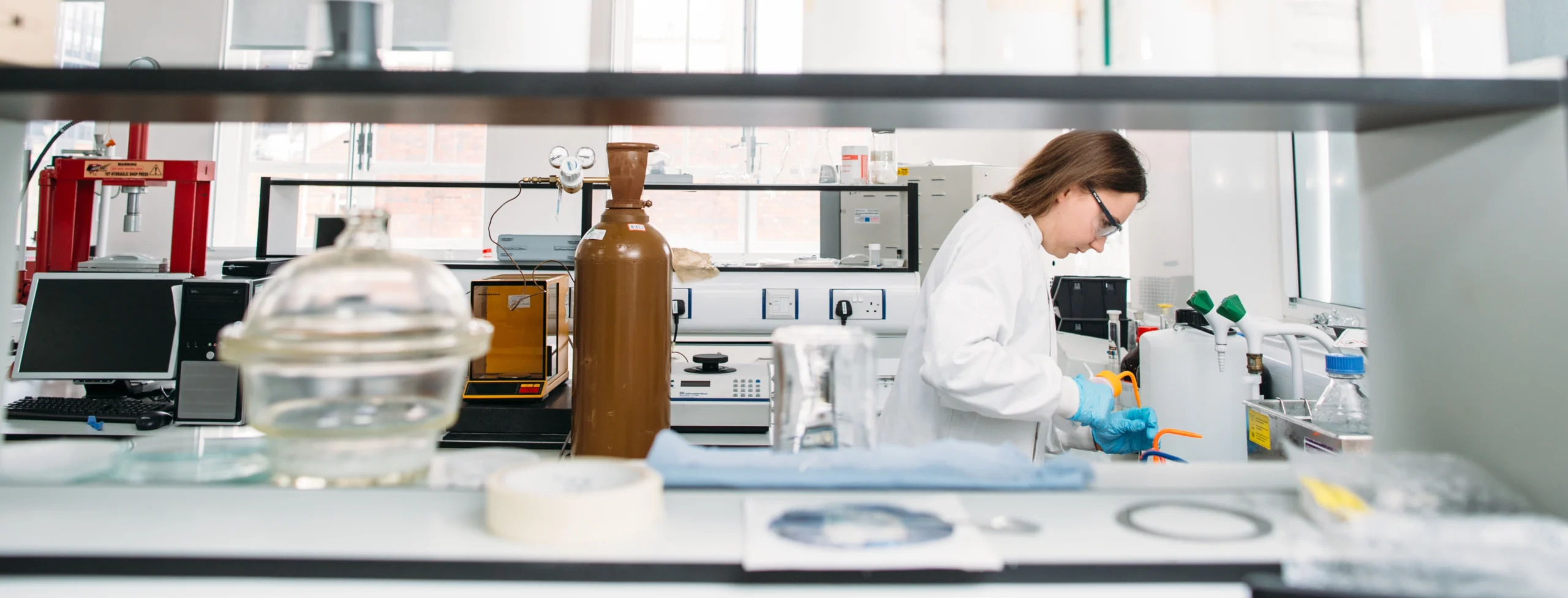 Scientist in white coat working at laboratory bench with beakers, bottles, and equipment in modern research facility.