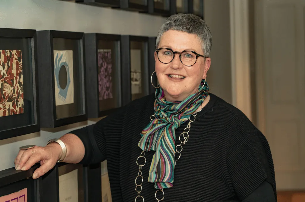 Dr Eleanor Kennedy smiling at the camera, wearing glasses and a colourful scarf, standing next to framed artwork.