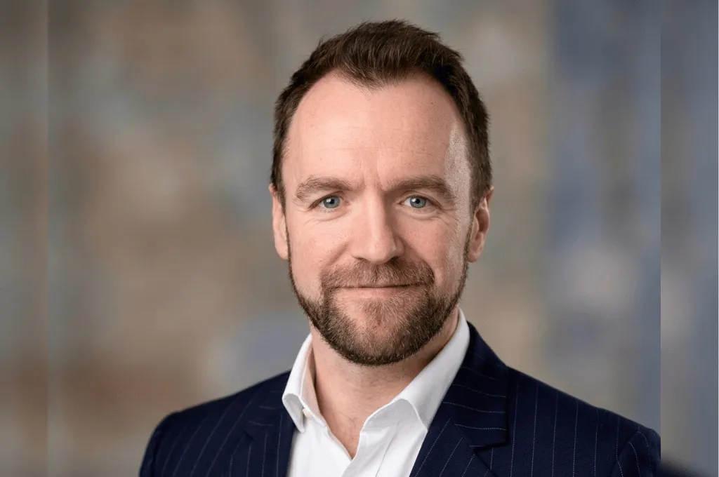 Professional headshot of Sam Tillotson in dark pinstriped blazer and white shirt against blurred background.