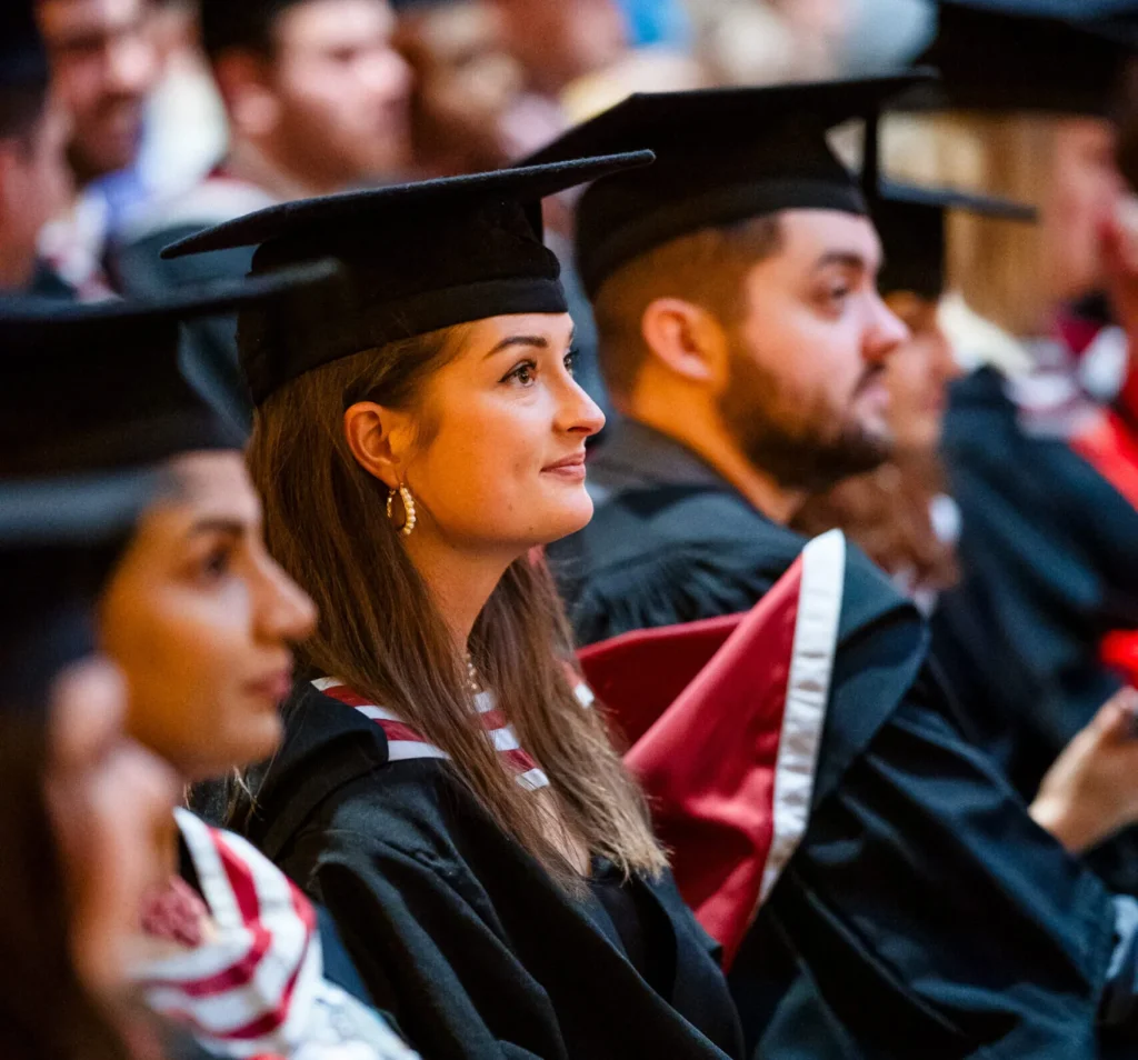Suz Mendus in academic robes and mortarboards at a graduation ceremony, seated among other students.