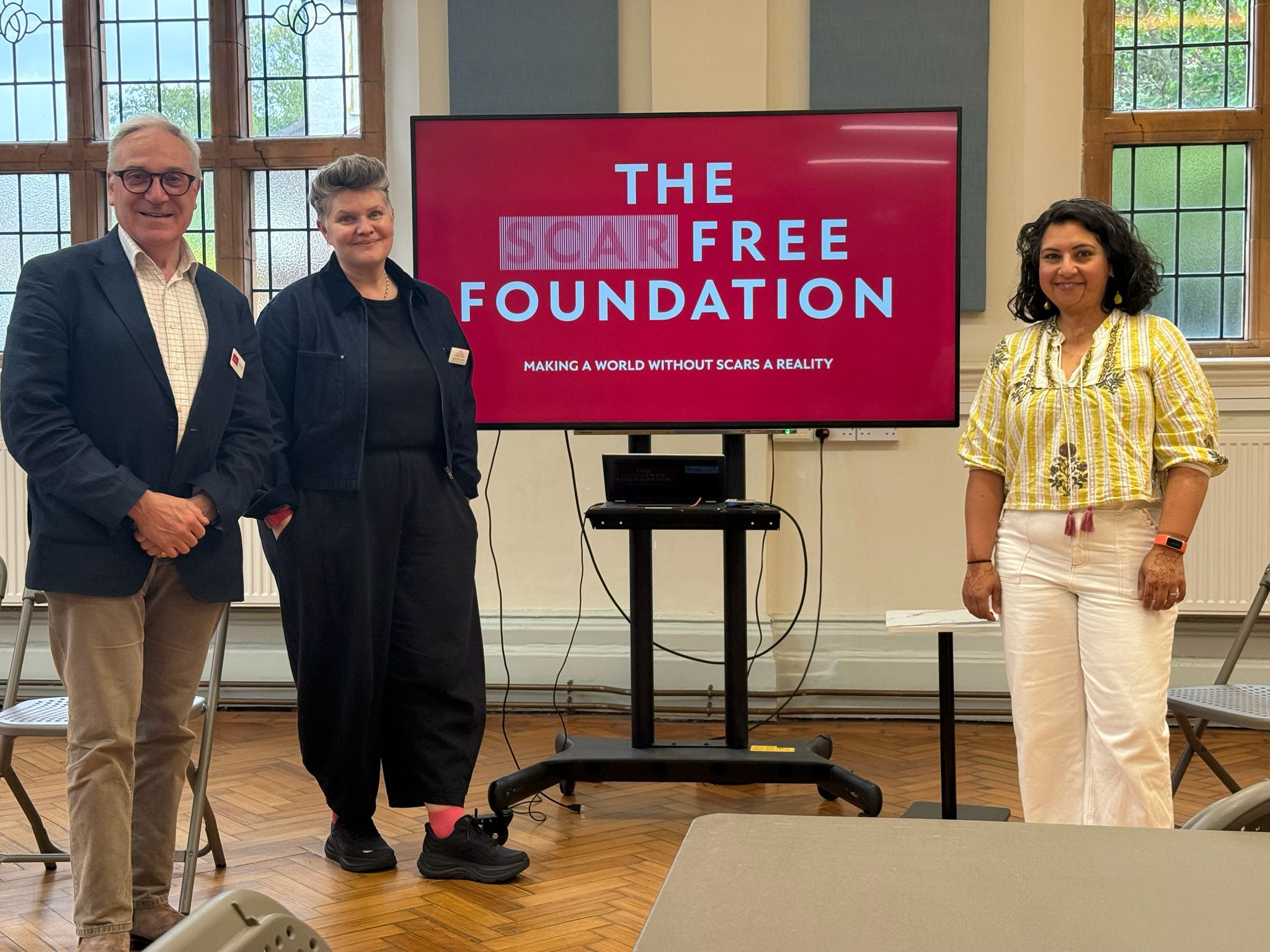 Richard, Char and Hemani stand in a line with a screen between Hemani and Charlotte. Hemani wears a yellow and cream shirt and trouser outfit, Charlotte wears a black top and trousers, and Richard wears beige trousers, a pink shirt, and a blazer. The screen has a red display and reads 'The Scar Free Foundation' in white. The three stand in a brightly lit room. From the left: Lieutenant General Richard Nugee, Charlotte Coates (Head of Research at The Scar Free Foundation), Hemani Modasia-Shah (a GP, and Scar Free Foundation Trustee and Ambassador)