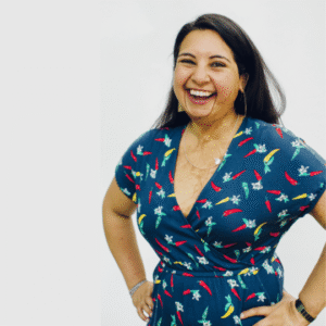 Dr Hemani Modasia-Shah wears a blue short dress patterned with green and red birds, as she smiles, standing with her hands on her hips