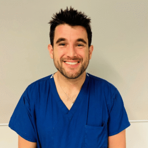 Mr Simon Filson, Consultant Plastic Surgeon at Evelina London Children's Hospital, stands against a white hospital wall wearing dark blue short sleeved scrubs and smiles 