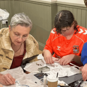 Andrea Jester and a volunteer wearing a bright orange football top work on garments at a crafts table. Andrew wears a red top and a beige jacket.