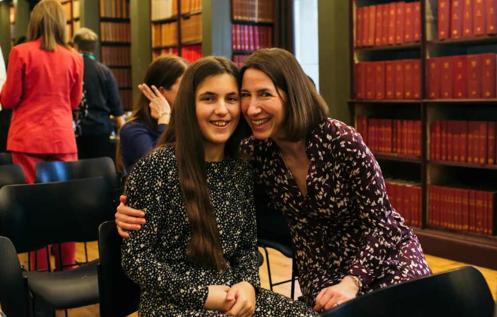 Two women smiling together in a library setting with bookshelves in the background.