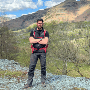 Ben Gallagher, an ex-British Army Special Forces Veteran stands on a stoney path with mountains, trees and grass in the background. Ben wears grey walking boots, trouser, t-shirt and a red backpack. He stands with his arms folded and smiles at the camera