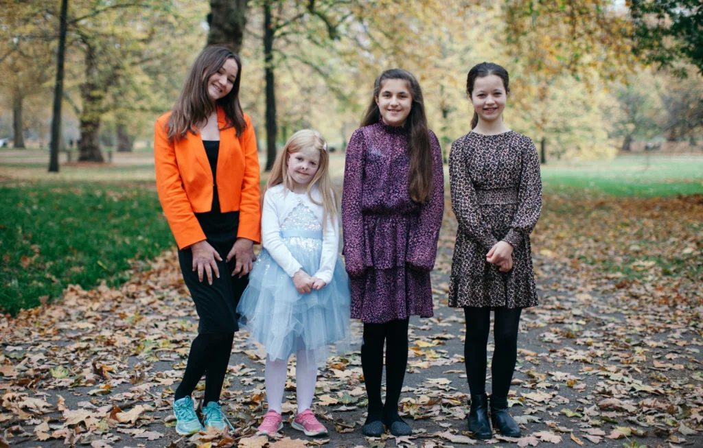 Four young girls standing together outdoors on an autumn path lined with fallen leaves and trees.