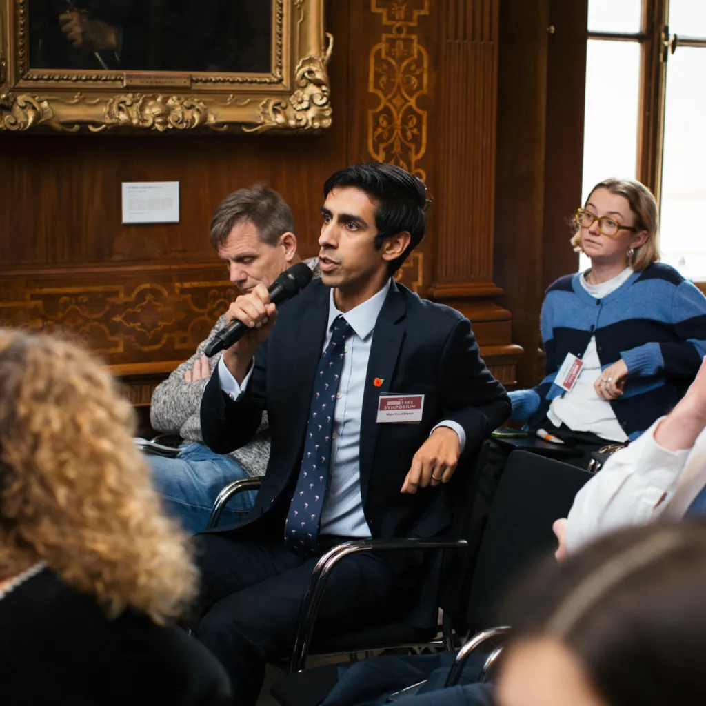 Man in business blazer speaking at panel discussion in ornate wooden room with seated participants.
