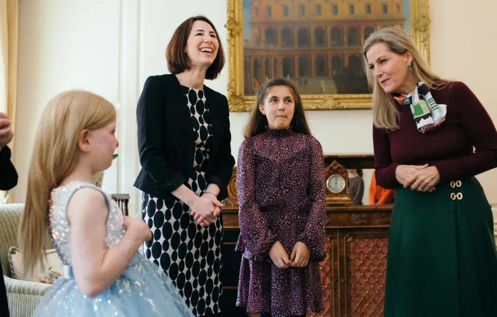 Two women and two girls in formal attire conversing in an elegant room with gold-framed artwork and furniture.