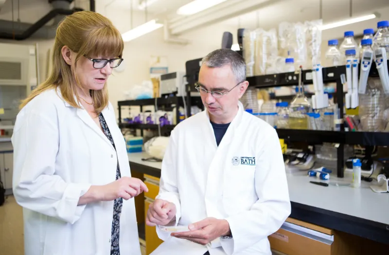 Two researchers in white lab coats examining a sample in a laboratory setting with equipment and bottles visible.