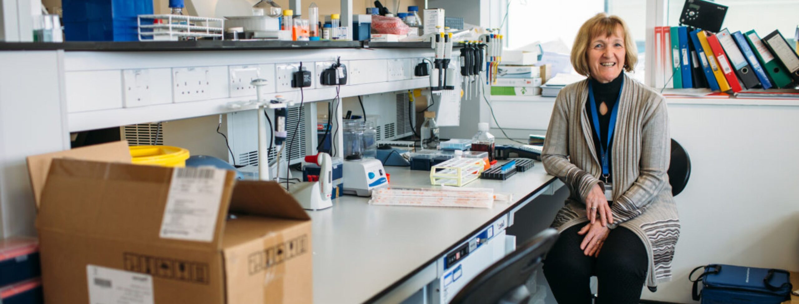 Professor Janet Lord wears a black top and grey cardigan as she sits at a desk covered in research equipment. Janet smiles at the camera.