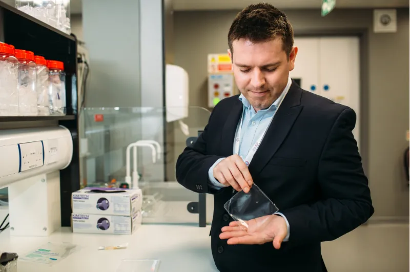 Man in business attire examining a dark medical device in a laboratory setting with equipment visible.