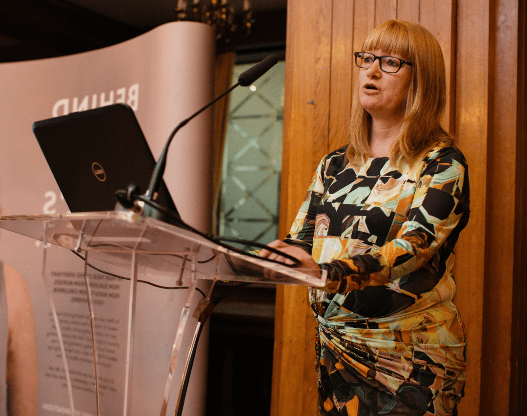 Professor Amber Young stands at a theatre lectern to deliver a talk at a Scar Free Foundation event. Amber had red shoulder length hair, wears black glasses, and a yellow, gold, and black patterned dress
