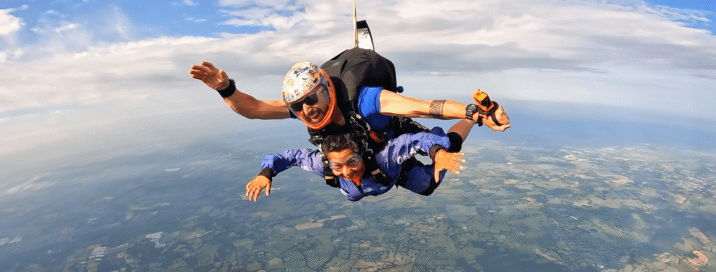 Hemani wears a blue jumpsuit and is attached to a skydive instructor wearing a parachute. They sail through the sky at 12,000 feet and there are patchwork green fields far below them and blue sky and clouds above them.