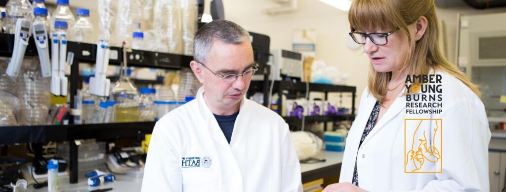 Professor Amber Young stands inside a laboratory wearing a white lab coat. Amber has shoulder length red hair, a fringe, and wears black glasses. She discusses something with a seated colleague. He also wears black glasses, has short grey hair, and wears a white laboratory coat too.