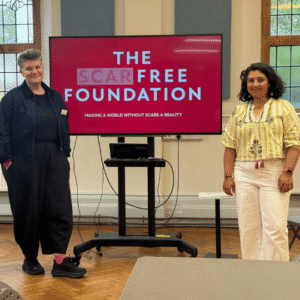 Hemani and Charlotte stand in an oak-floored library room in front of a TV screen reading 'scar free foundation'. Hemani stands to the right and wears a yellow short-sleeved blouse and white trousers. Charlotte stands to the left and wears a black shirt and trousers. 