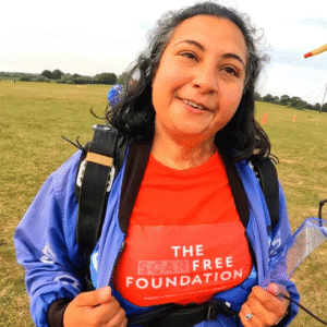 Hemani wears a blue jumpsuit opened to reveal a red 'The Scar Free Foundation' t-shirt. She is standing in the green field where she landed her skydive.
