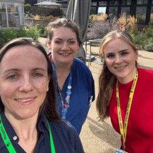 Olivia Hartrick wears a green lanyard and blue shirt on a sunny day in Horatio's Garden in Stoke Mandeville hospital. To Olivia's right wearing a bright blue shirt is Charlotte Bowman, a Paediatric Burns nurse, and Alex Stanley, one of the plastic surgery core trainees, who wears a red shirt and a yellow lanyard. Olivia takes a selfie as they all smile at the camera. 