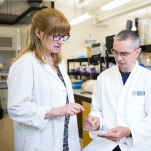 Professor Amber Young stands inside a laboratory wearing a white lab coat. Amber has shoulder length red hair, a fringe, and wears black glasses. She discusses something with a seated colleague. He also wears black glasses, has short grey hair, and wears a white laboratory coat too. 