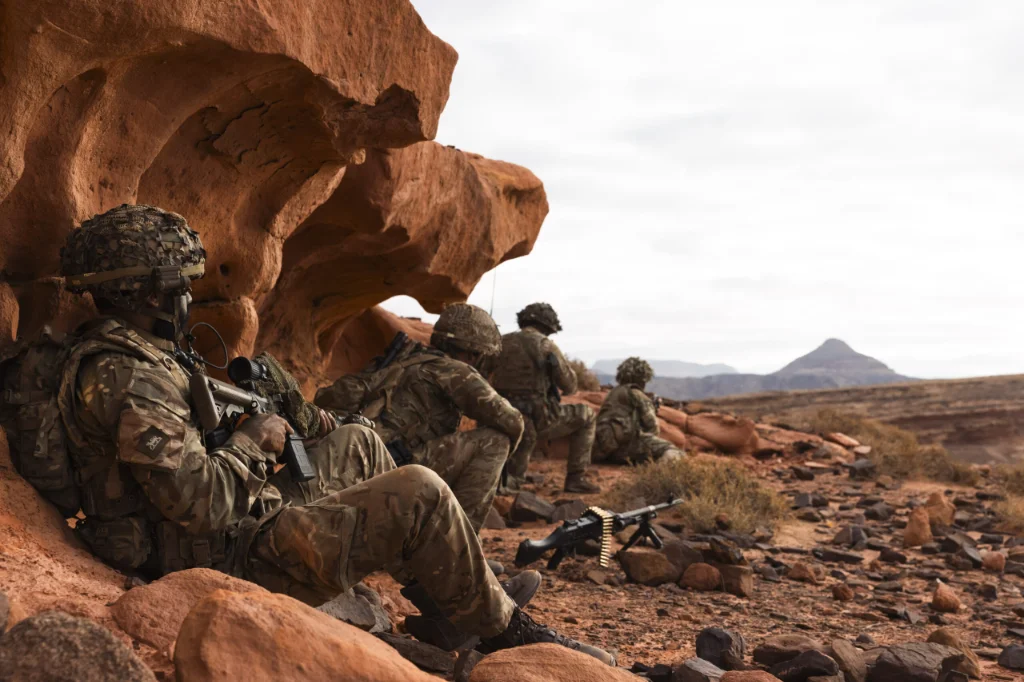 Four soldiers in camouflage carry a soldier in a stretcher to a dark green medical vehicle where another soldier waits.