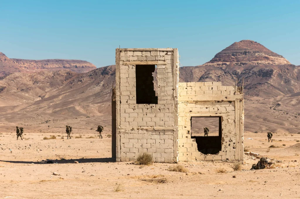 A derelict sandstone brick building in the desert, with mountainous terrain behind. Five soldiers patrol in the distance.