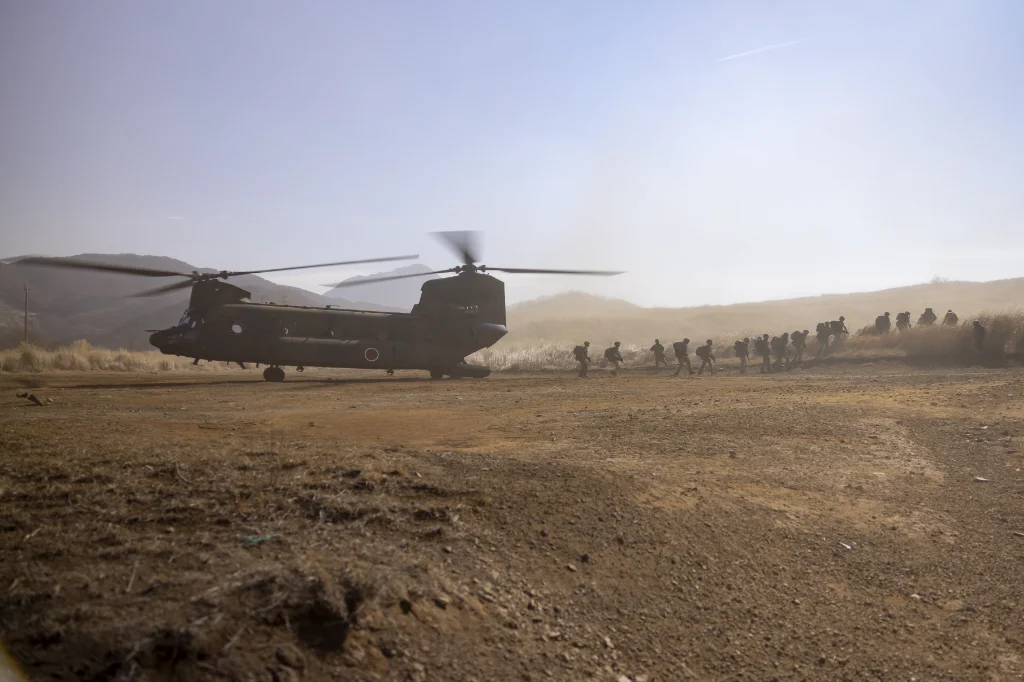 A CH47 double-motored helicopter landed on a dusty plain, with a long line of soldiers leaving the aircraft next to it.