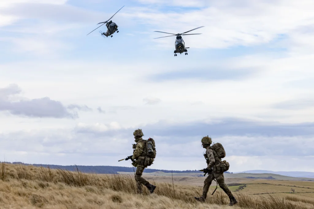 Two armed soldiers wearing camouflage trek across grassy Welsh mountains as two chinook helicopters fly overhead.