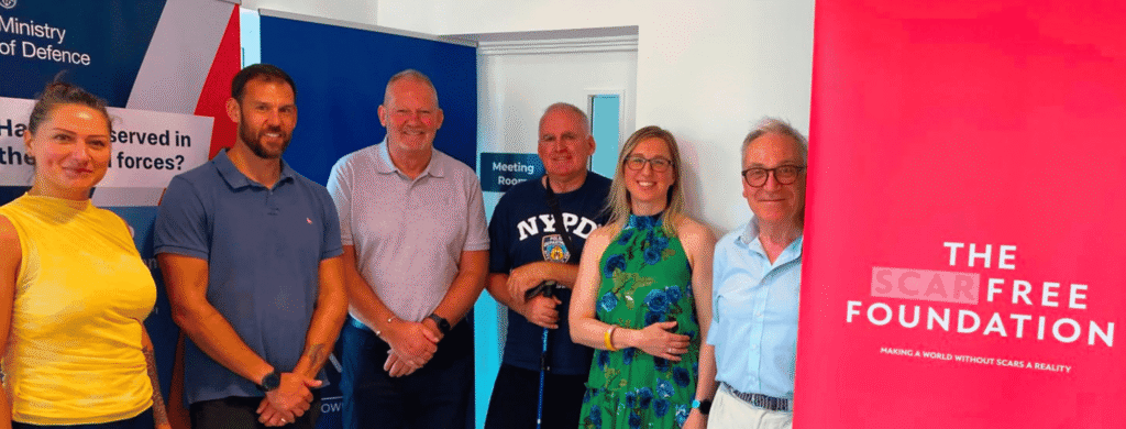 Smiling attendees at The Scar Free Foundation's second community event, in association with Royal British Veteran's Enterprise. Six people stand in a line in front of a Ministry of Defence banner and a Scar Free Foundation banner. From left to right: Anna Bratu, Ben Gallagher, Steve Hammond, an attendee Veteran, Kristina Stiles, Richard Nudge