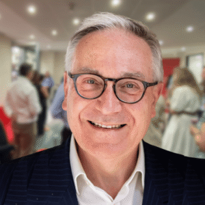 Lt Gen (Rtd) Richard Nugee smiles at the camera. There is a blurred superimposed image behind him of the Summer Reception event, showing people in a brightly lit office room. In the foreground, Richard wears glasses, a white shirt, and a blue suit.