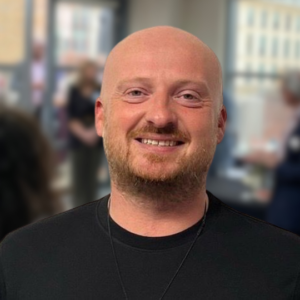 Iwan Roberts-Mok wears a dark blue round neck jumper and smiles at the camera. In the background, a room full of people at the event is blurred.