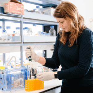 Dr Beck Richardson, a biologist and researcher at the University of Bristol, wears a black jumper and white latex gloves as she works at a lab. She stands over a bench and uses a pipette to drop fluid into a bright yellow vial.