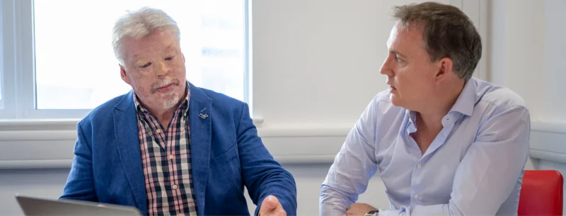 SImon Weston CBE in mid blue jacket with laptop in meeting with man in light blue shirt perched on a red char in an office.