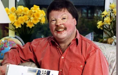 Simon Weston CBE in ox red shirt sitting on a cream sofa with daffodils and a world globe behind him.