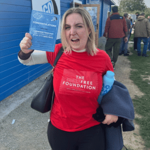 India Gale, Scar Free Foundation Ambassador, celebrates her skydive - she holds a blue certificate in the air, wears a red Scar Free Foundation t-shirt and stands in front of a blue wooden building.