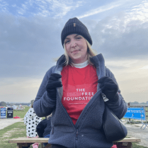 India Gale, a Scar Free Foundation Ambassador, stands in a field with a blue sky behind her. India wears a red Scar Free Foundation t-shirt, black puffer coat, dark blue gloves and a dark blue wooly hat. She has shoulder length blonde hair.