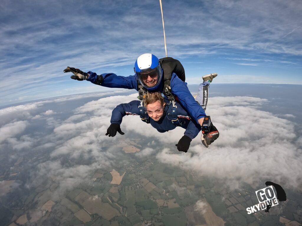 India Gale soars through the sky strapped to an instructor as she does her skydive. They both wear blue jumpsuits and black harness. The curve of the earth can be seen below them.